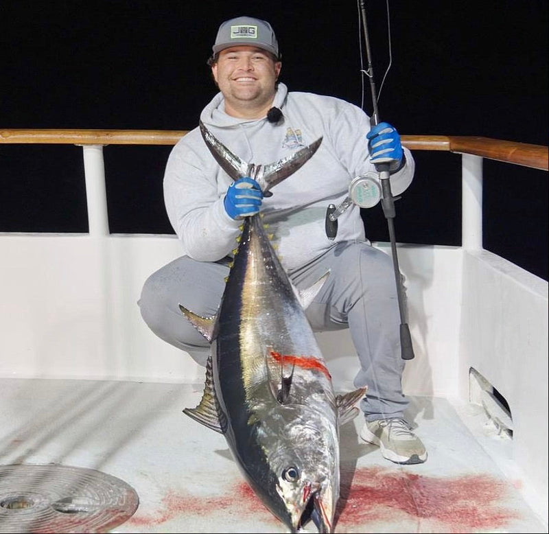 Angler in a JYG cap holding a bluefin tuna by the tail on a boat deck at night