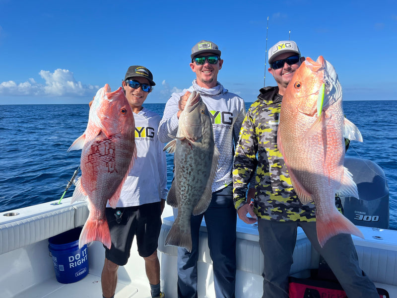 Three anglers in JYG shirts holding two red snapper and a gag grouper on a boat offshore