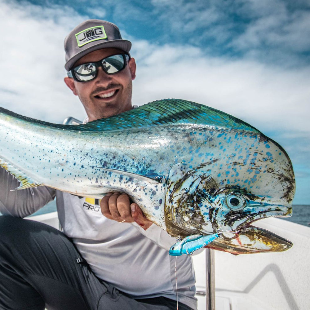 A man wearing a gray cap with a visible 'BIG' logo and sunglasses sits on a boat, holding a large blue and silver mahi-mahi fish under his arm against an ocean and cloudy sky background.
